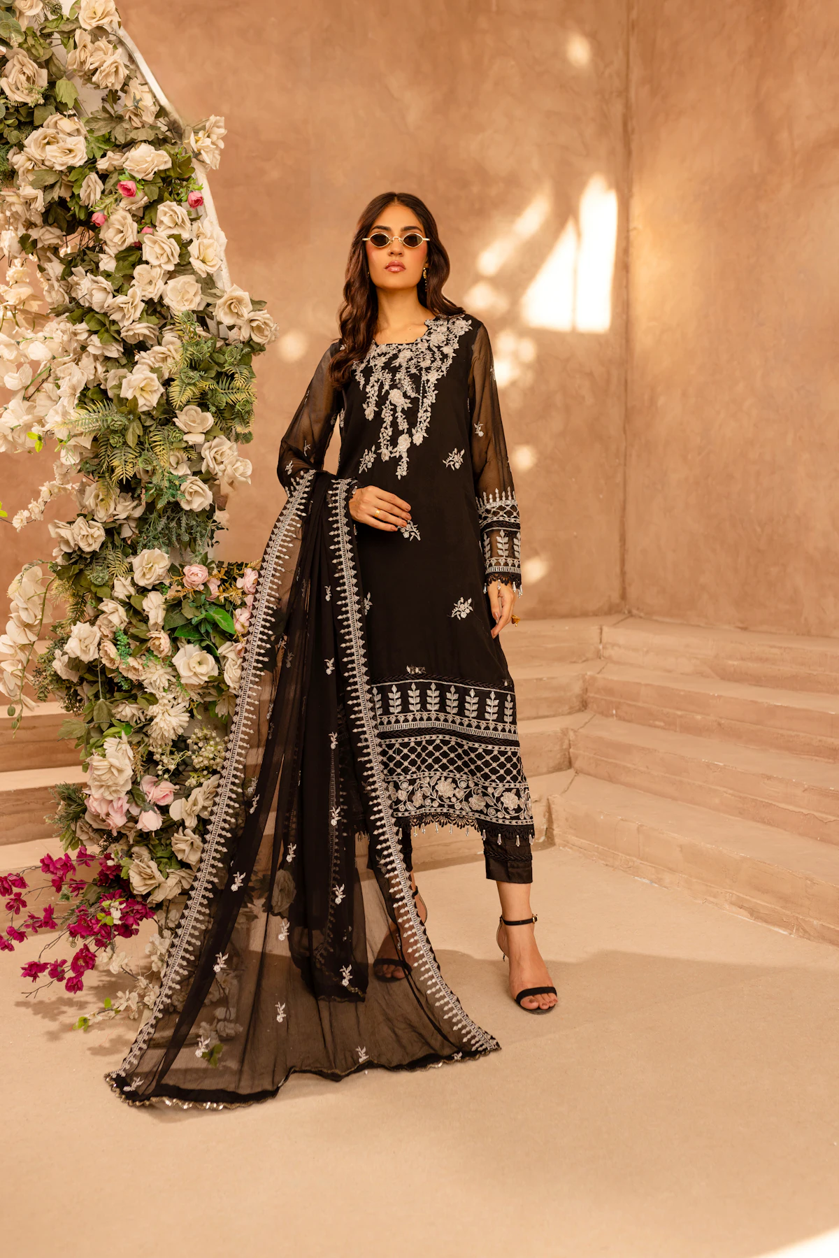 A woman in sunglasses wearing a black net formal with white embroidery and a long embroidered dupatta.