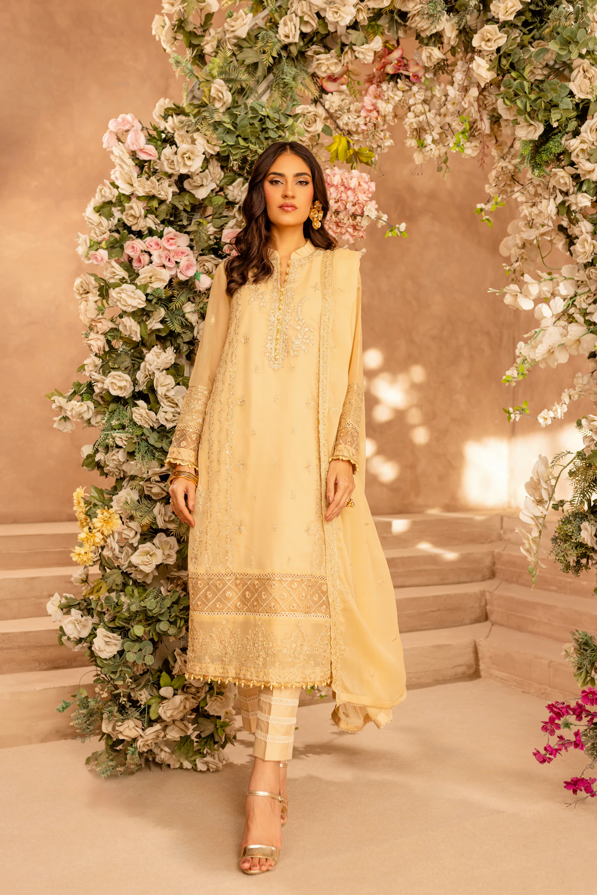 A woman in a pale butter yellow lawn suit with white embroidery and a matching dupatta, standing under a flower arch.