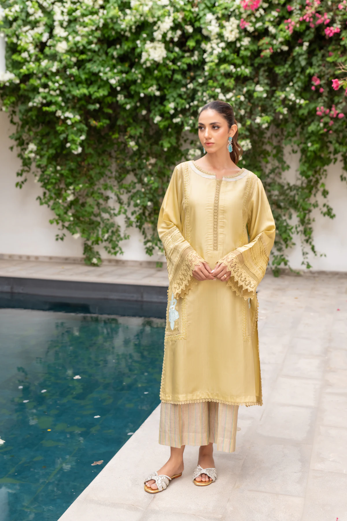A woman in an antique gold lawn suit with an embroidered neckline, standing by a pool under hanging vines.
