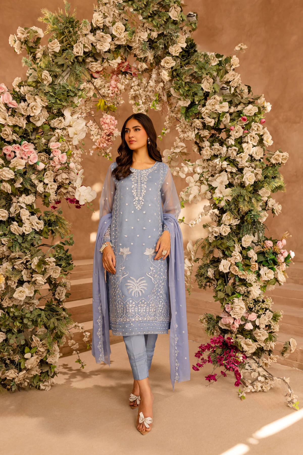 A woman in an ice blue organza formal with tonal embroidery and a long dupatta, under a flower arch.