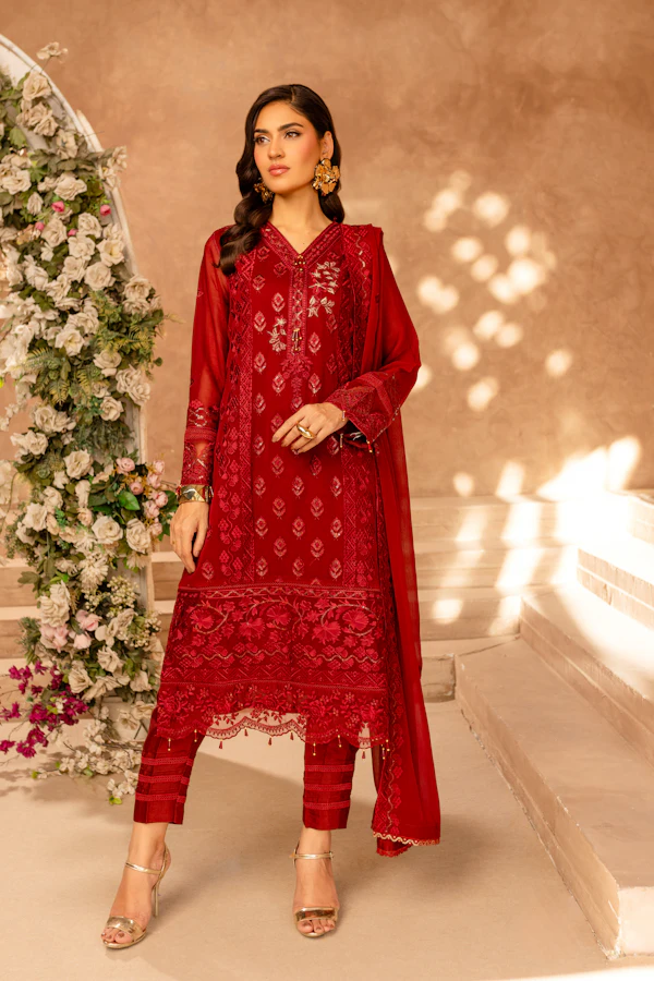 A woman in a deep red formal suit with gold zari embroidery and a red chiffon dupatta, by a flower arch.